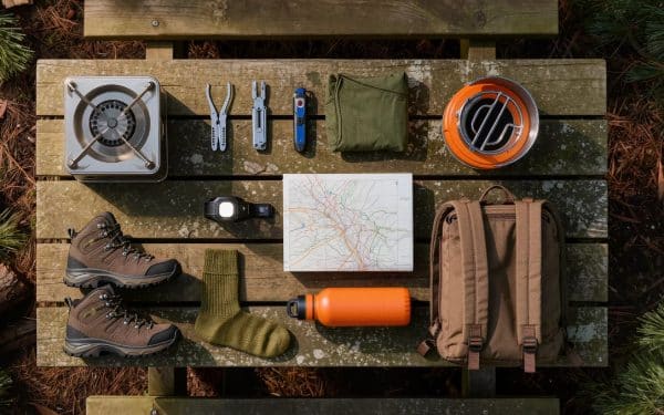 Outdoor adventure gifts flat lay with hiking boots, camping stove, multi-tool, headlamp, water bottle, and backpack on weathered picnic table