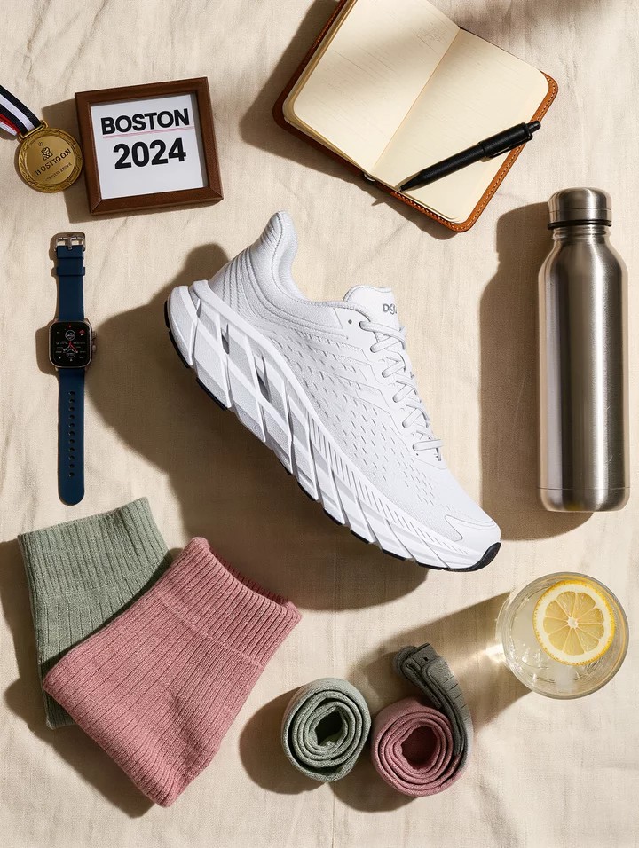 Gifts for runners who have everything, flat-lay of Hoka running shoe, GPS watch, folded run socks, framed race bib, finisher medal, stainless water bottle, training journal, and compression sleeves on cream linen